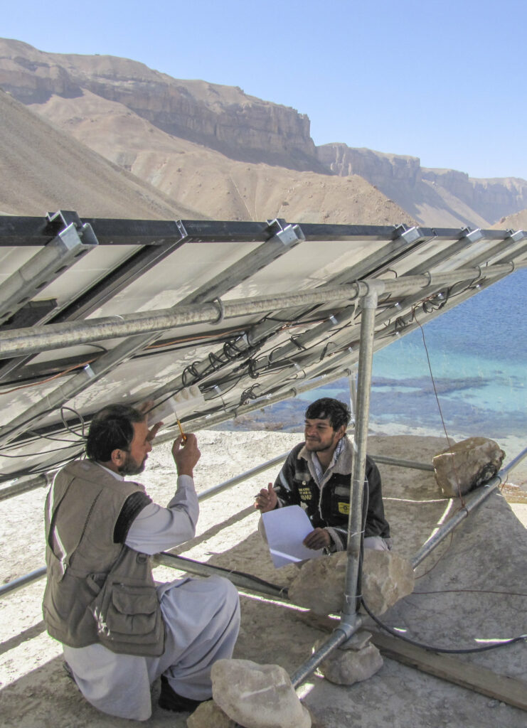 two men work underneath a solar panel in the bright sunshine by a lake