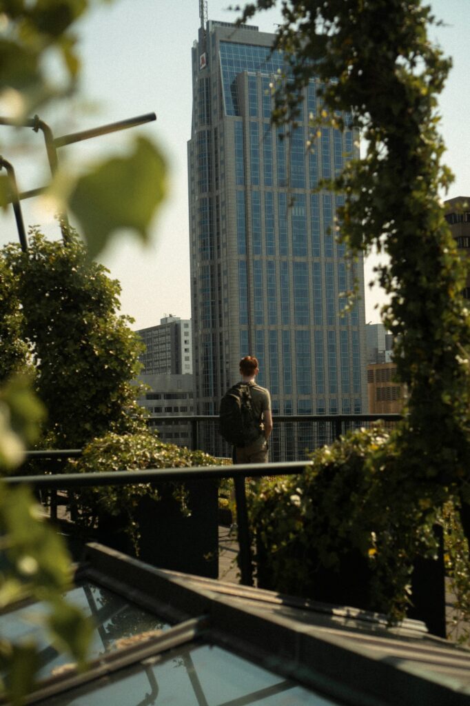 man stands and looks at tall building. He is framed by green trees